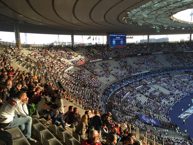 Large crowd in a stadium watching a soccer game with a stage, flags, poles, and a screen in the background, identified as the Allianz Arena in Munich, Germany.
