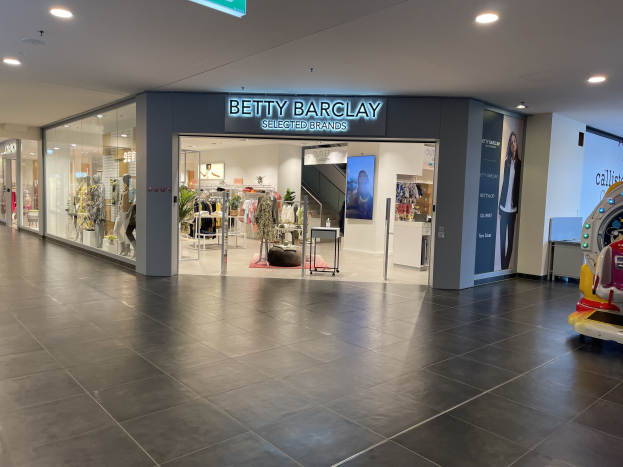Interior of a Betty Barclay store in a shopping mall displaying dresses on mannequins, clothing on hangers, tables, plants, a toy vehicle on the right, and a sign reading "Betty Barclay Selected Brands" at the top.
