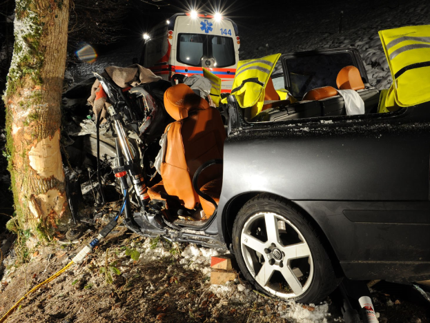 Ein verunfalltes Auto am Straßenrand auf einer schneebedeckten Straße mit einem im Hintergrund sichtbaren Krankenwagen.