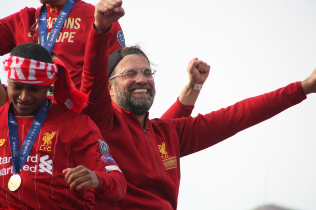 Liverpool-Trainer Jürgen Klopp feiert in roter Jacke mit seinen Spielern den Sieg in der UEFA Champions League, umgeben von zwei medaillentragenden Personen und dem Himmel im Hintergrund.