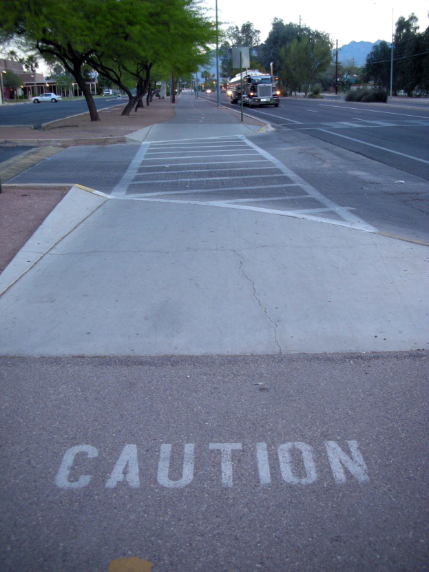 Stadtstraße mit fahrenden Fahrzeugen, ein Gehweg mit einem Warnschild neben einem Baum, Strommasten, Straßenlaternen, Bauzäune, Grünflächen, entfernte Gebäude und Berge unter einem klaren blauen Himmel.
