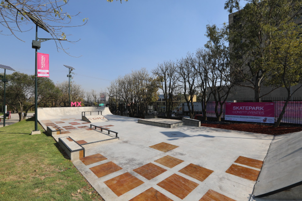 Ein Skatepark mit einer zentralen Rampe, umgeben von Bäumen, Gras, Laternen, Bannern und einem Zaun, mit Gebäuden und einem klaren blauen Himmel im Hintergrund.