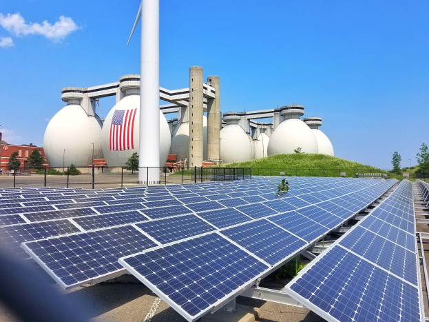 Solarpanels auf dem Dach eines Gebäudes mit einer Flagge, umgeben von einem Zaun, Gras, Pflanzen, Bäumen und anderen Objekten, mit zusätzlichen Gebäuden und einem bewölkten Himmel im Hintergrund.