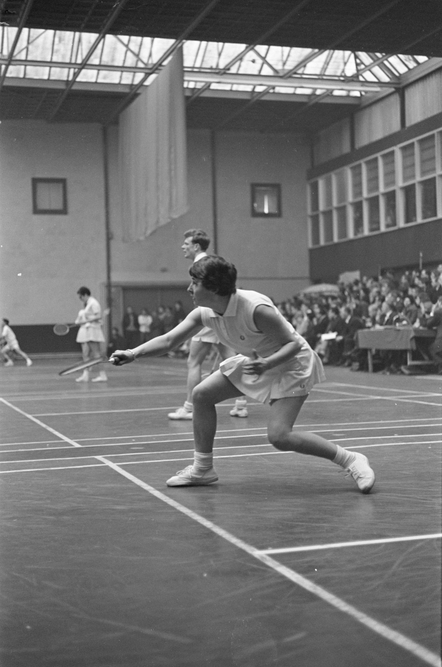 Schwarzes und weißes Foto von zwei Frauen, die Badminton auf einem Court spielen, jeweils einen Schläger haltend, mit Zuschauern rechts und einer fensterumrandeten Wand im Hintergrund.