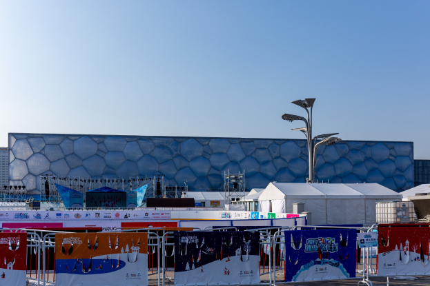 Olympiastadion in Sotschi, Russland, mit Vordergrund-Elementen wie Bannern, Geländern, Zelten, Laternenmasten und Gebäuden vor einem klaren blauen Himmel.