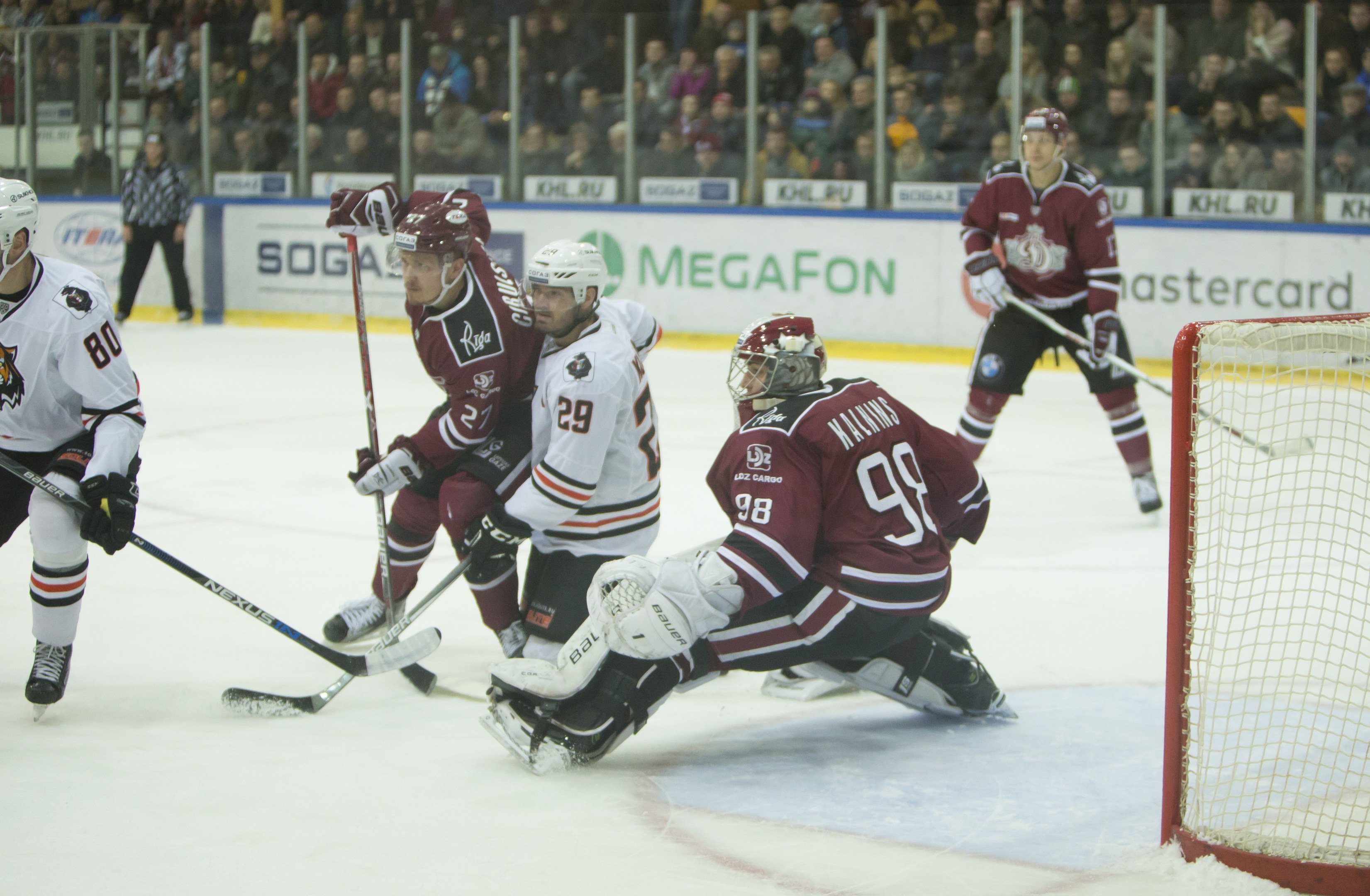 Gruppe von Menschen, die Hockey auf einem Eisstadion spielen, mit Torpfosten auf der rechten Seite, tragen Helme und halten Hockey-Schläger, Zuschauer auf Tribüne mit Bannern im Hintergrund.