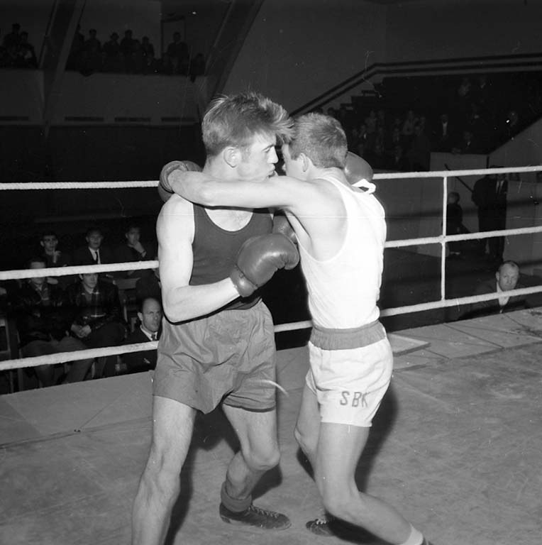 Zwei Boxer kämpfen im Ring, umgeben von sitzenden und stehenden Zuschauern, auf einem Schwarz-Weiß-Bild.