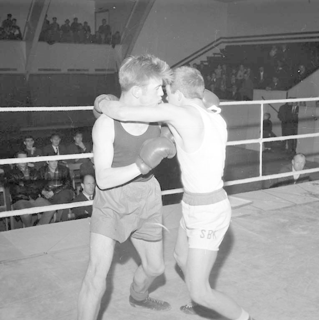 Zwei Boxer kämpfen im Ring, umgeben von sitzenden und stehenden Zuschauern, auf einem Schwarz-Weiß-Bild.