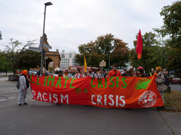 A group of people holding a "Climate Crisis is a Crisis" banner walking down a tree-lined street with vehicles, buildings, and a clear blue sky in the background.