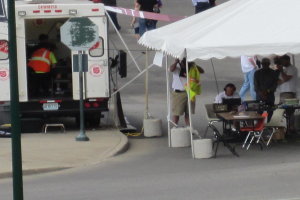 Ein Rettungswagen steht links von einer Straße, während rechts ein Zelt viele Menschen schützt, mit einem Tisch und einem Stuhl in der Nähe.