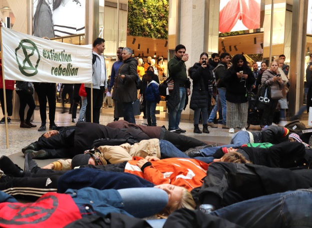 Menschen liegen auf dem Boden vor einer Menge mit Handys und Taschen, mit einem Banner im Vordergrund und Protestschildern, Puppen und Säulen im Hintergrund.