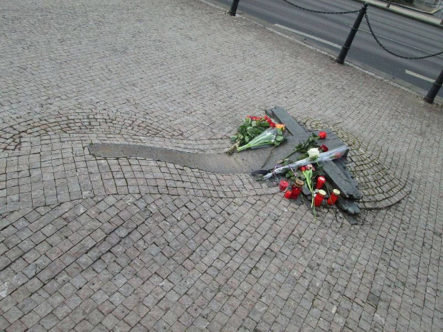 Gedenkstätte mit Blumen, Kerzen und verstreuten Sträußen am Straßenrand, umgeben von Bäumen, Gebäuden und Fahrzeugen mit Kettenpfählen.