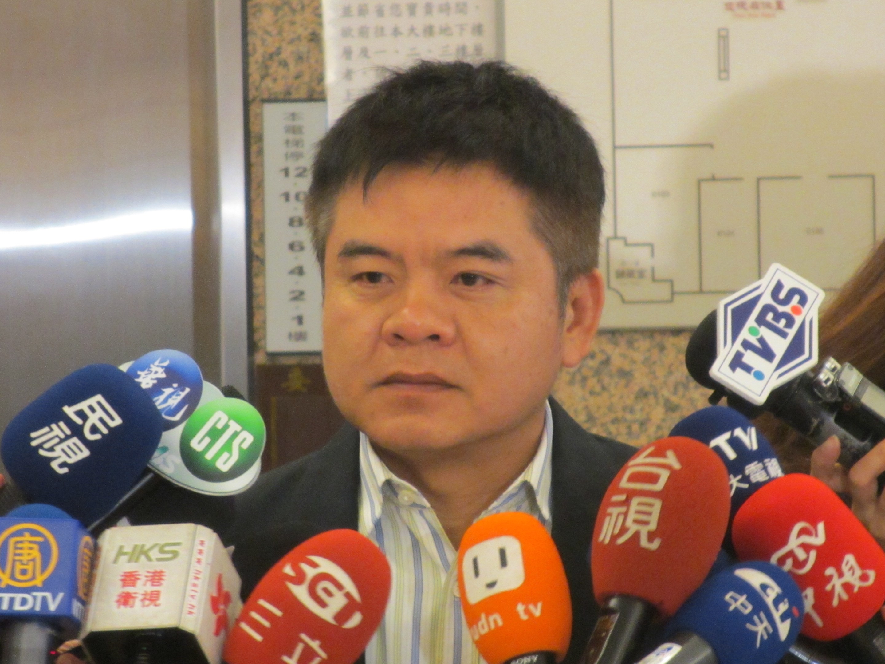 Ein Mann im Anzug und Krawatte spricht vor einer Wand mit einer Tafel zu Reportern, während eine Frau rechts eine Kamera hält.