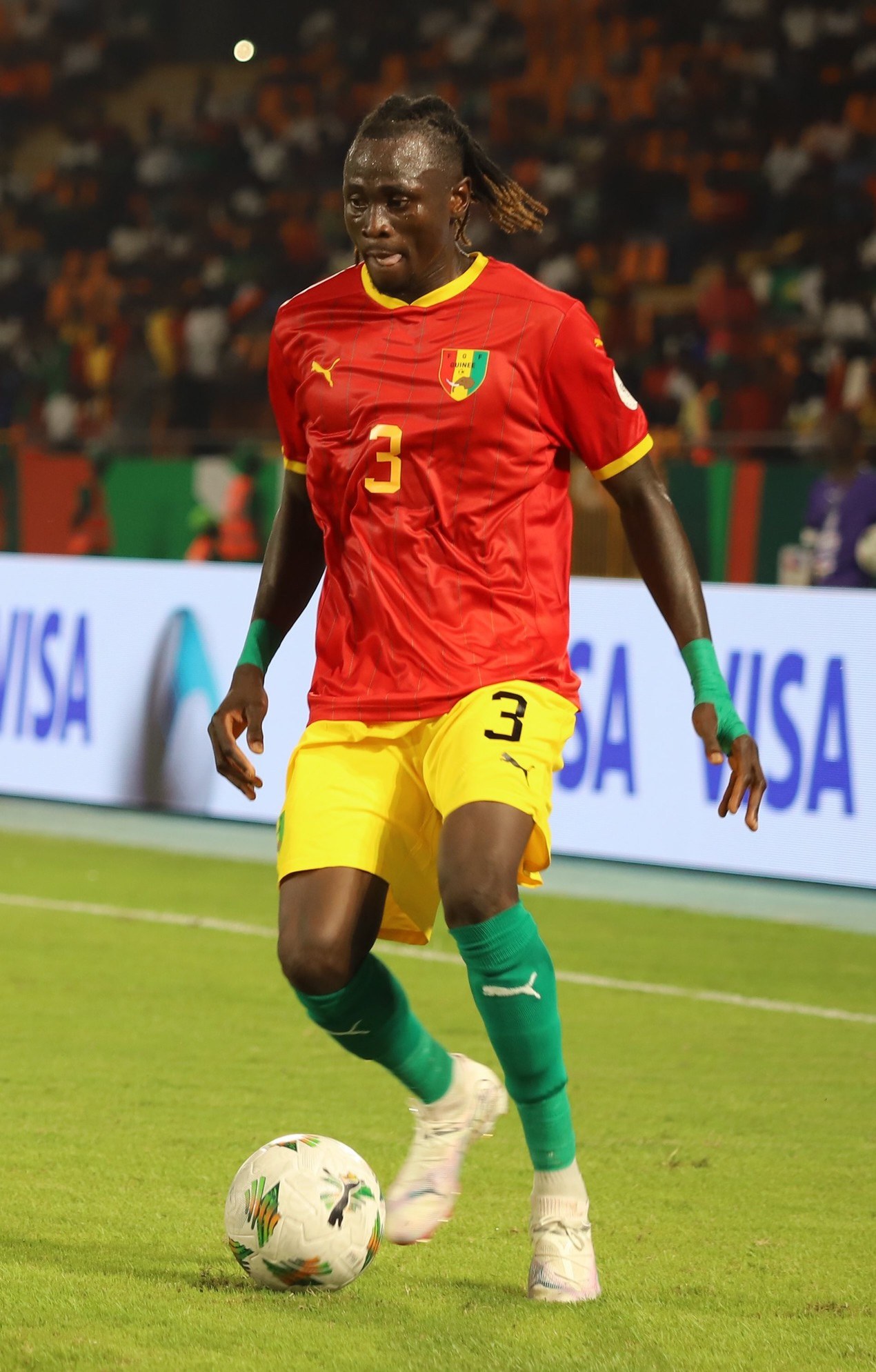 Ein Fussballspieler in rot-gelber Uniform, der auf einem Feld mit Zuschauern in den Rängen und Werbetafeln im Hintergrund einen Ball tritt.
