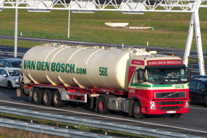 Ein großer weißer, roter und grüner Tanklastwagen fährt auf einer Autobahn, umgeben von anderen Autos, mit einer Begrenzung am Straßenrand, Gras im Hintergrund und einer Eisenrahmenkonstruktion weiter hinten.