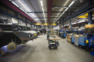 Ein großes Lagergebäude voller Maschinen, einschließlich Fahrzeugen auf der linken Seite, Transportwagen mit Gegenständen, Holzboxen und anderen Gegenständen auf der rechten Seite sowie Lampen an der Decke.