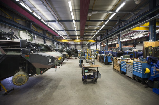 Ein großes Lagergebäude voller Maschinen, einschließlich Fahrzeugen auf der linken Seite, Transportwagen mit Gegenständen, Holzboxen und anderen Gegenständen auf der rechten Seite sowie Lampen an der Decke.