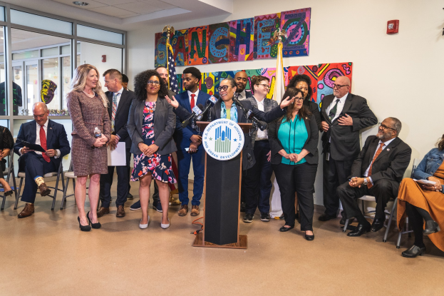 Gruppe von Menschen vor einem Rednerpult mit Mikrofon und einer Tafel mit der Aufschrift "Sancho Stadtrat tritt dem Stadtrat bei", einige sitzen auf Stühlen und andere stehen, mit Fahnen und einem Feuerlöscher an der Rückwand.
