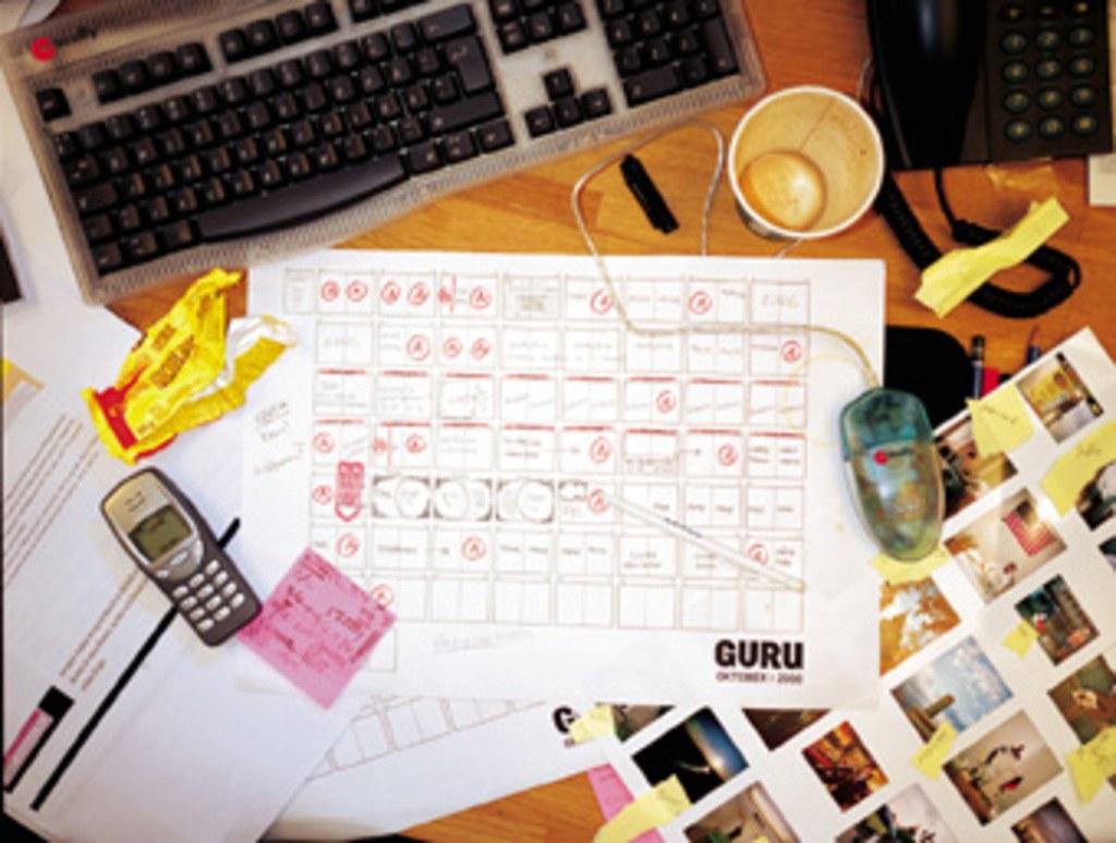 Ein unordentlicher Schreibtisch mit Papieren, einem Computer, einem Telefon, Tassen und einem Mobiltelefon.