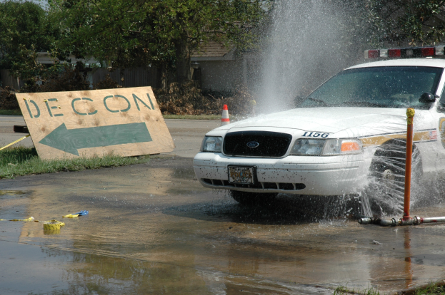 Ein Polizeiwagen fährt durch eine Pfütze, während verstreute Gegenstände, darunter eine Tafel mit Text und ein Verkehrskegel, auf dem Boden in der Nähe von Bäumen, Gebäuden und einem klaren blauen Himmel liegen.