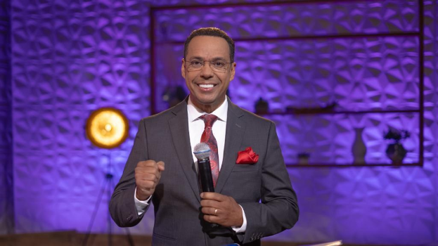 Stephen Colbert, Moderator von 'The Daily Show with Trevor Noah', steht auf einer Bühne und hält ein Mikrofon, trägt einen Anzug, eine Krawatte und eine Brille, mit einer Lampe und einem Ständer hinter ihm.