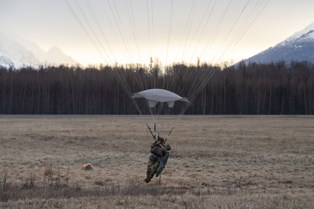 Person paragliding over a grassy field with snow-covered mountains in the background, wearing a helmet under a bright blue sky.