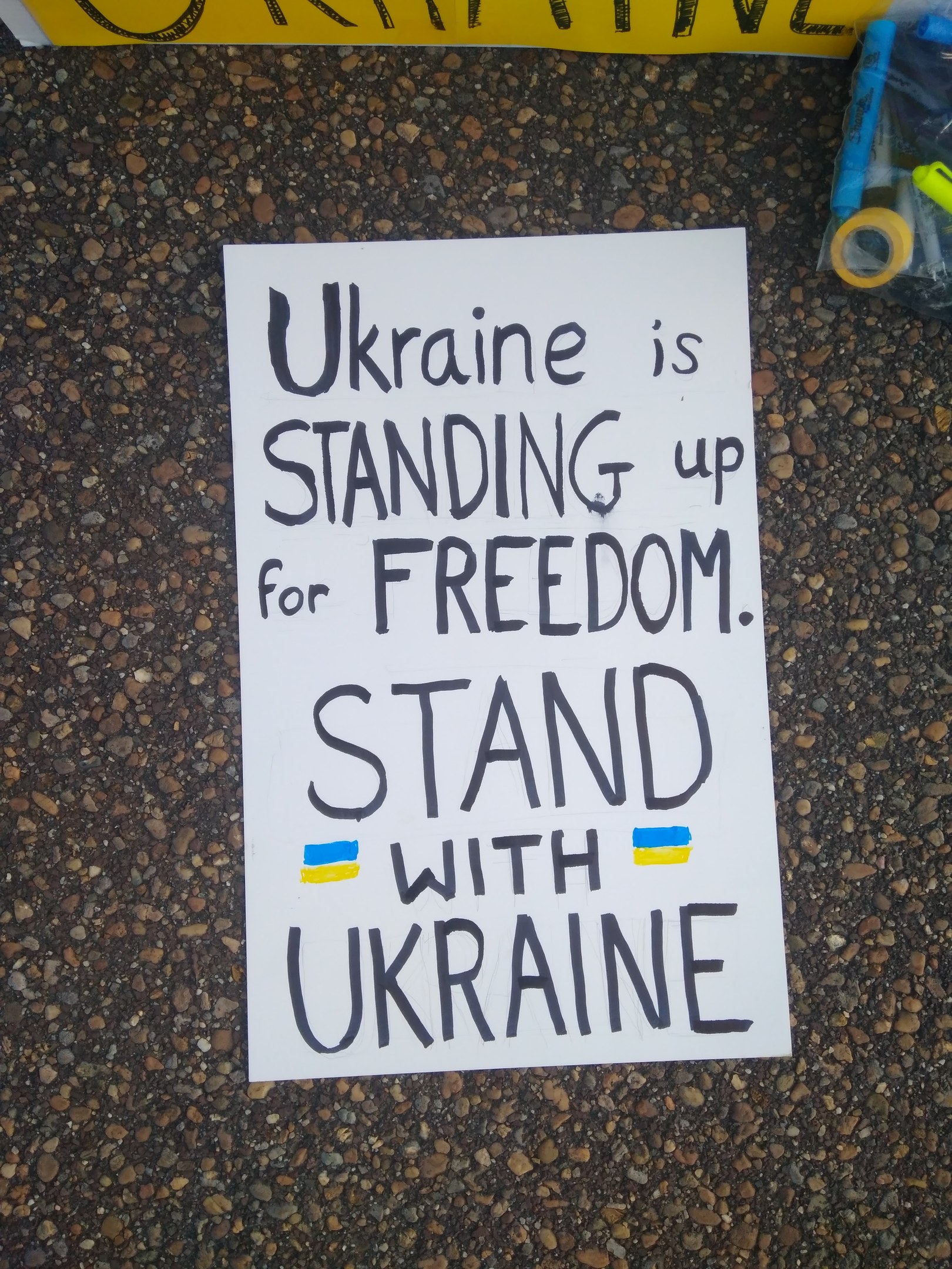 Weißer Zettel mit "Ukraine steht für Freiheit, steht mit Ukraine" Text auf dem Boden vor gelbem Banner, mit Plastikabdeckung mit Gegenständen rechts.