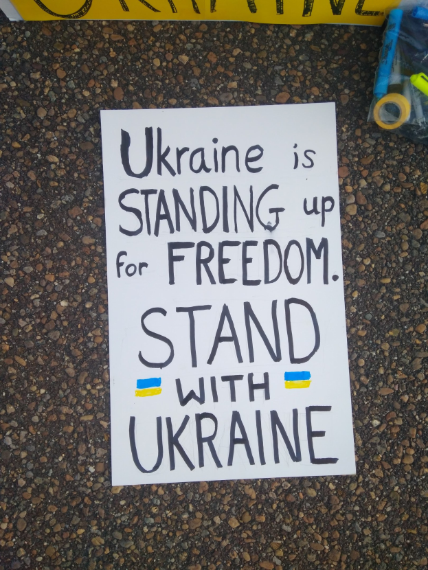 Weißer Zettel mit "Ukraine steht für Freiheit, steht mit Ukraine" Text auf dem Boden vor gelbem Banner, mit Plastikabdeckung mit Gegenständen rechts.