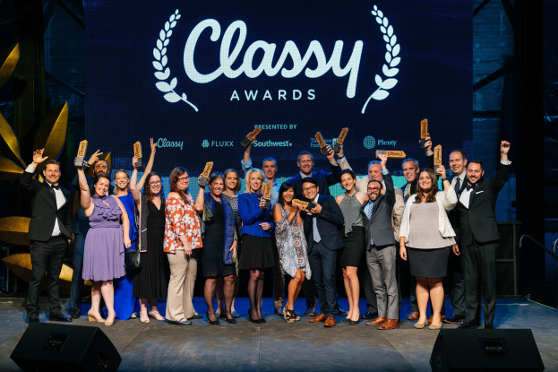 Gruppe von Menschen auf einer Bühne, die Auszeichnungen halten, mit Lautsprechern darunter und einem Bildschirm im Hintergrund mit der Aufschrift "Classy Awards 2019."