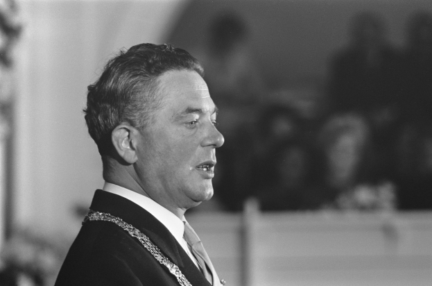 Ein Mann im Anzug und Krawatte steht an einem Podium mit einem Mikrofon und spricht zu einer unscharfen Menge, die wie ein Politiker wirkt.