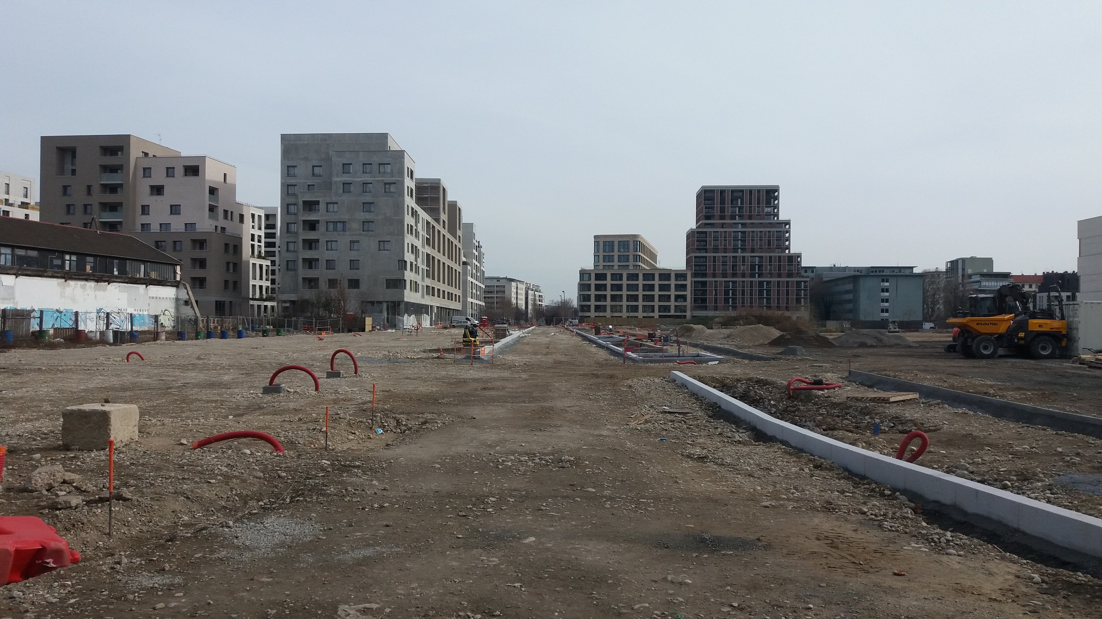 Baustelle mit schwerer Machinery im Vordergrund, Gebäude und Bäume im Hintergrund unter klarem Himmel.