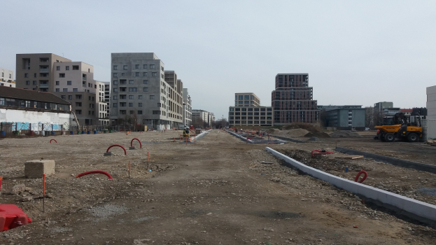 Baustelle mit schwerer Machinery im Vordergrund, Gebäude und Bäume im Hintergrund unter klarem Himmel.