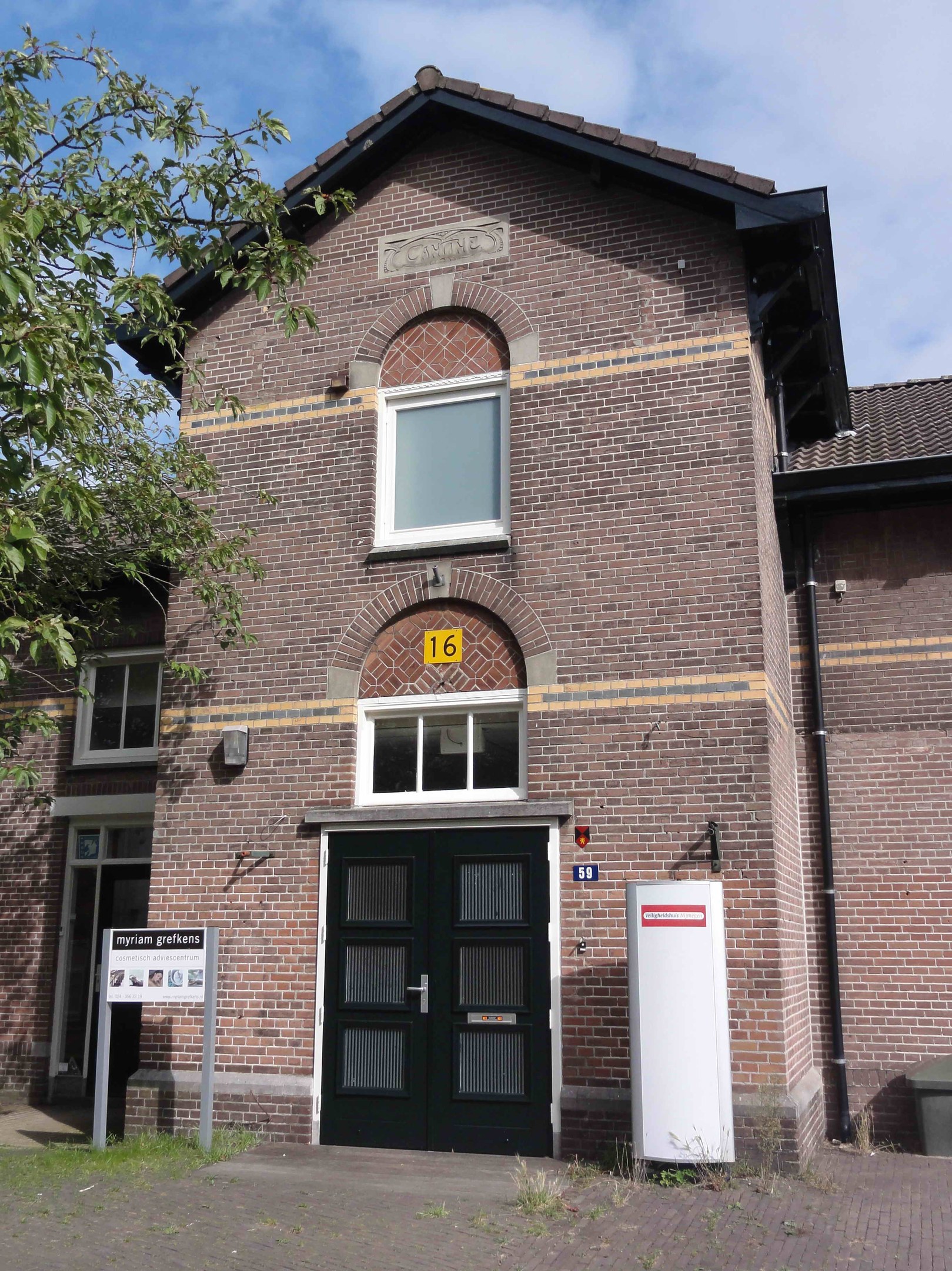 Ziegelbau mit grüner Tür und Fenstern, ein Schild, ein Müllcontainer, Topfpflanzen, ein Baum und ein bewölkter Himmel.