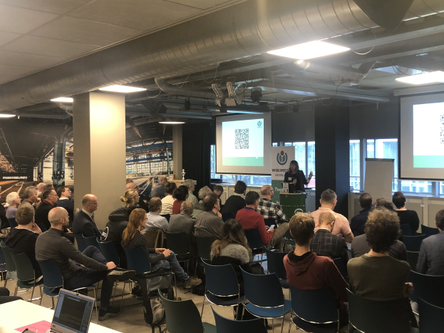 Eine große Gruppe von Menschen sitzt auf Stühlen und schaut auf eine Projektionsfläche, während ein Redner neben einem Mikrofon auf einem Lautsprecherständer steht, in einem Raum mit Text an einer Tafel, Säulen, Fenstern, Deckenleuchten und einer Decke.