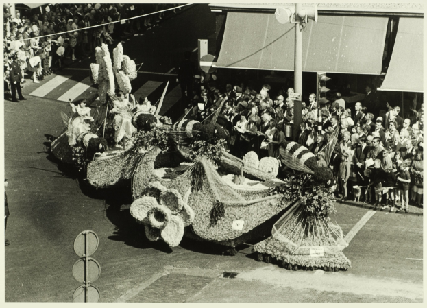 Ein Schwarz-Weiß-Foto eines mit Blumen geschmückten Umzugswagens, mit einer Gruppe von Menschen auf der Straße und einem Mast im Hintergrund.