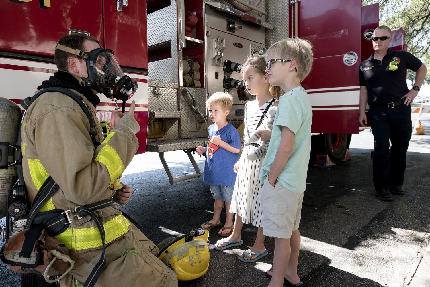 Feuerwehrmann mit Gasmasken spricht zu Kindern vor einem Feuerwehrauto mit Bäumen und blauem Himmel im Hintergrund.