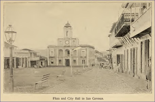 Ein Schwarz-Weiß-Foto des Rathauses von San German mit umliegenden Gebäuden, Straßenlaternen, Bänken, Menschen und einem Uhrenturm im Hintergrund sowie Text am unteren Rand.