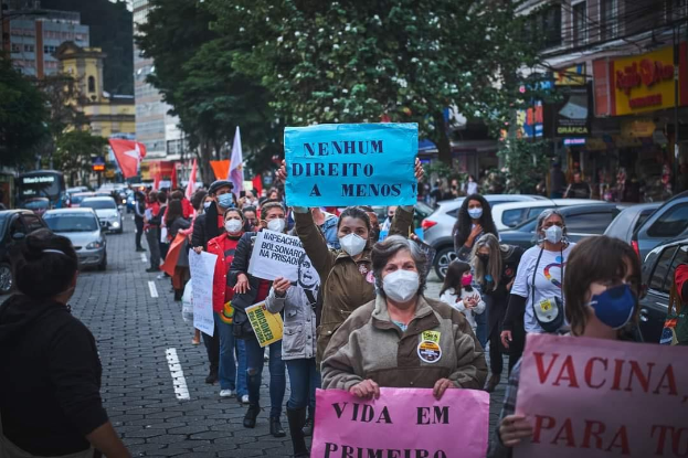 Group of people wearing masks and holding signs in a street protest against a government vaccine ban in Brazil, surrounded by vehicles, trees, buildings, and text boards.