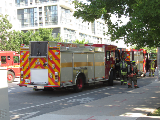 Eine Gruppe von Feuerwehrautos auf einer Straße geparkt, mit Menschen in Helmen und Taschen auf dem Bürgersteig, Gras auf der rechten Seite, Bäume und Gebäude im Hintergrund unter einem klaren blauen Himmel.