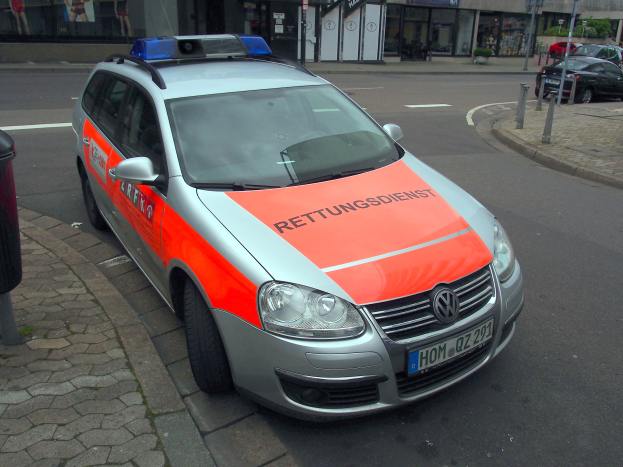 Polizeiauto auf dem Seitenstreifen neben einem Müllcontainer auf dem Gehweg geparkt, mit Gebäuden und Bäumen im Hintergrund.