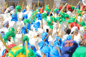 Eine große Gruppe von Menschen in farbenfrohen Kost√∞men marschiert eine Straße entlang, jeder hält einen Stock, mit einem Gebäude im Hintergrund.