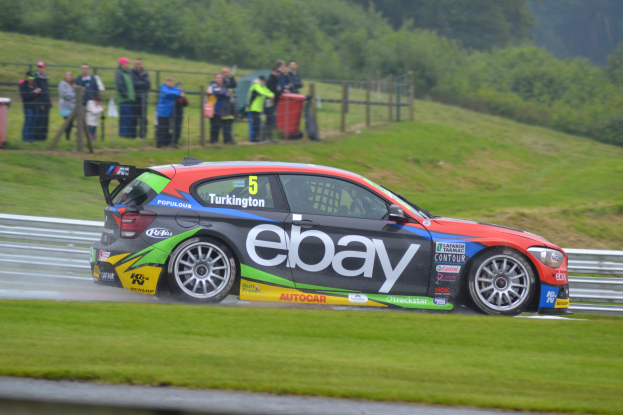 Ein Auto fährt auf einer Rennstrecke mit Rauch aus dem Auspuff, umgeben von Gras, einem Zaun, Mülltonnen, Menschen, Bäumen und einem klaren blauen Himmel.