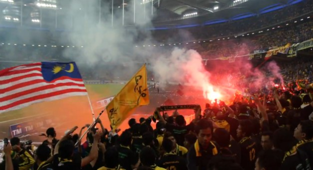 Eine große Menschenmenge in einem Stadion mit Fackeln, Fahnen und Rauch, mit Schildern, die 'Malaysia vs Malaysia Live Streaming' anzeigen.