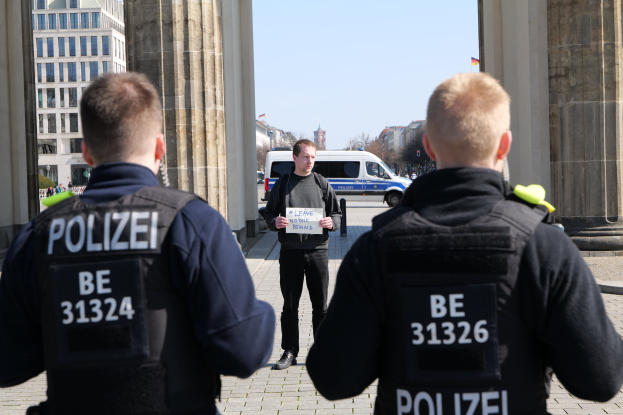 Drei Polizisten stehen vor einem Gebäude, wobei ein Mann in der Mitte ein Papier hält, flankiert von Säulen, während Fahrzeuge, Menschen, Bäume und Gebäude den Hintergrund unter dem Himmel füllen.