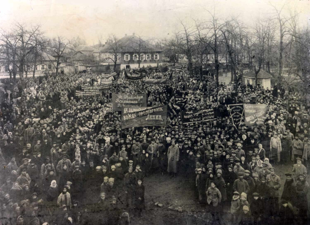 Black and white image of a large crowd holding banners in front of a building, with trees and windows visible in the background.