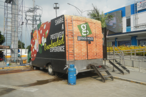 Ein Lkw steht vor einem Gebäude mit einer Tafel, auf der "Teilen Sie ein positiv überladenes Erlebnis" steht, mit Stufen, die zum Lkw führen, einem Zylinder auf dem Boden, Geländern, elektrischen Polen mit Drähten, Laternenpfählen, Bäumen und einem bewölkten Himmel im Hintergrund.