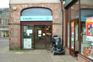 Eine Apotheke mit einem vor ihr geparkten Fahrzeug und einem Gebäude links im Bild.