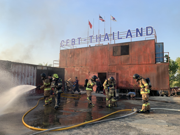 Feuerwehrleute in Helmen sprühen Wasser auf ein Gebäude mit Text, mit Flaggen, Bäumen und einem klaren Himmel im Hintergrund.