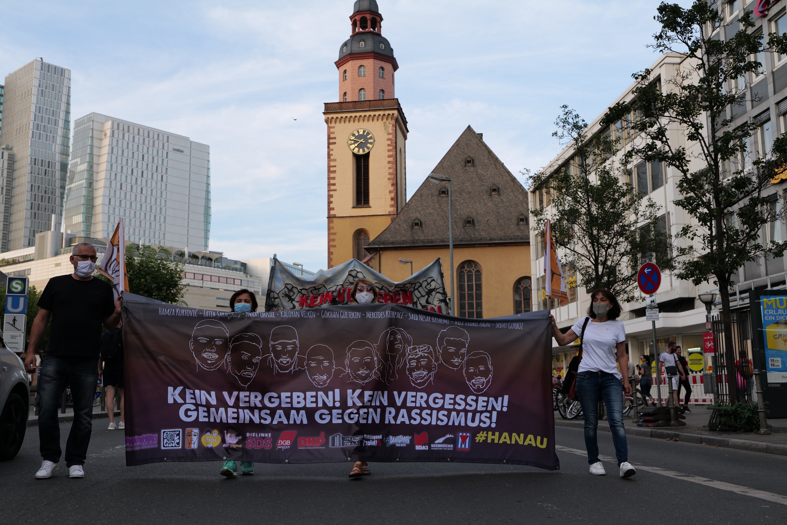 Eine Gruppe von Menschen in Masken, die eine Transparente mit Text und Bildern tragen, öffnet sich auf einer Straße, mit einem geparkten Auto auf der linken Seite, Gebäude, Bäume, Schilder, Mäste, ein Uhrenturm und einen klaren blauen Himmel im Hintergrund.