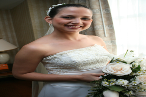 Eine strahlende Braut mit einem Bouquet in der Hand vor einem Vorhang und einer Wand im Hintergrund und einer Lampe auf einem Tisch links.